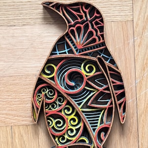 Può includere: Scultura di pinguino in legno a strati con disegni intricati e colorati. Il pinguino presenta strati di rosso, giallo, blu e nero, creando un effetto 3D. L'opera d'arte è ambientata su uno sfondo di legno.