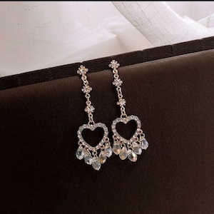 May include: A pair of gold-tone dangle earrings with a heart-shaped design. The hearts are outlined with clear rhinestones and have a dangling row of clear teardrop-shaped crystals.