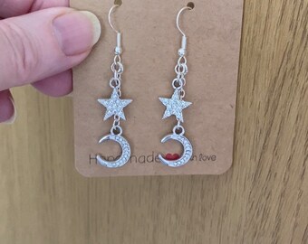 May include: Silver earrings with a crescent moon and star charm. The charms are decorated with clear rhinestones. The earrings are on a brown card with the text "Handmade with love".