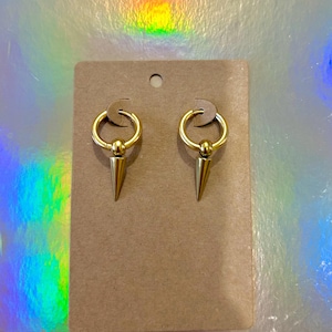 May include: A pair of gold-toned earrings with a hoop and a pointed spike. The earrings are displayed on a brown card.