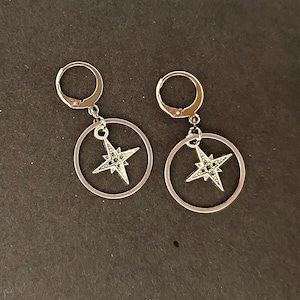 May include: A pair of silver earrings with a star design. Each earring features a small hoop with a dangling star charm inside a larger circle.