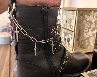 May include: Black leather combat boot with silver chain and sword charms. The boot features a side zipper, studded accents, and buckle straps. A hand is holding the boot.