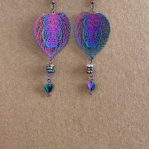 May include: A pair of iridescent owl earrings. Each earring has a leaf-shaped pendant with a cut-out owl design. Below the owl are three small, colourful beads and a heart-shaped charm. The earrings have a hook closure.