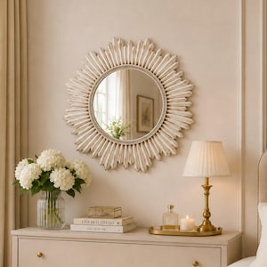 May include: A round sunburst mirror with a cream-colored frame hangs above a three-drawer dresser. On the dresser sits a glass vase filled with white hydrangeas, a stack of books, a small glass bottle, a candle, and a brass table lamp.