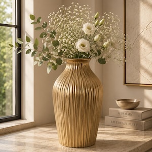 May include: A tall, gold-colored vase with a textured, vertical-lined design. The vase holds a bouquet of white flowers and green foliage. The vase is approximately 18 inches tall and sits on a light-colored surface.