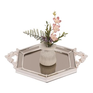 May include: A hexagonal mirrored tray with a white ornate frame and two decorative handles. Centered on the tray is a small, ribbed, off-white vase containing a bouquet of artificial pink flowers, green fern leaves, and small purple sprigs.