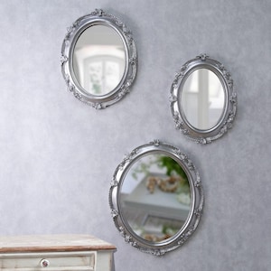 Può includere: Tre specchi ovali con cornici argentate ornate sono appesi a una parete grigio chiaro. Gli specchi riflettono l'ambiente circostante. Le cornici presentano dettagli floreali e a volute, aggiungendo un tocco di eleganza.