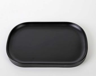 May include: A matte black rectangular tray with rounded corners and slightly raised edges. The tray features a smooth, solid-colored surface and a wide, flat base, providing a simple and functional design for holding various small items.