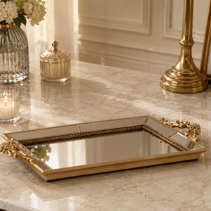 Op de afbeelding: Een rechthoekige, goudkleurige spiegeltray met sierlijke handgrepen. De tray heeft een spiegeloppervlak en een gouden frame. Een glazen kandelaar met een brandende kaars, een glazen pot en een vaas met witte bloemen zijn ook zichtbaar.