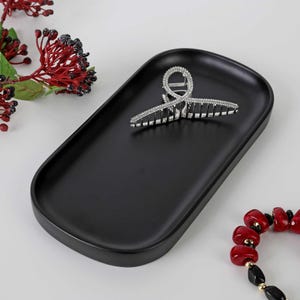 May include: A matte black oval tray holds a silver-toned hair claw clip encrusted with clear crystals. The tray is positioned near a cluster of red berries with green leaves and a bracelet featuring red and black beads with gold accents.