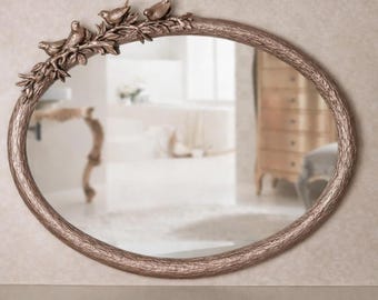 Op de afbeelding: Een ovale wandspiegel met een metalen bronskleurige lijst die is getextureerd als boomschors. Het linkerbovengedeelte van de lijst bevat een decoratief tak-element met drie gebeeldhouwde vogels die tussen bladeren en twijgen zitten.