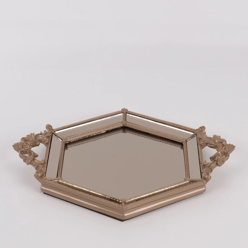 May include: A decorative tray with a mirrored surface and a gold-colored frame. The tray has a unique geometric shape with ornate handles on each side. The tray is designed to hold items and add a touch of elegance to any space.