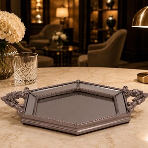 May include: A decorative, mirrored tray with a gray frame and ornate handles. The tray has a geometric shape and a reflective surface. The handles feature a detailed, floral design. The tray is displayed on a marble-like surface.