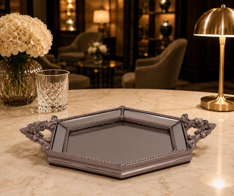 May include: A decorative, mirrored tray with a gray frame and ornate handles. The tray has a geometric shape and a reflective surface. The handles feature a detailed, floral design. The tray is displayed on a marble-like surface.
