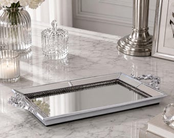 May include: A rectangular mirrored vanity tray with a silver-toned frame and decorative ornate handles on each side. The tray features a reflective glass surface bordered by a beaded trim, providing a polished and metallic aesthetic for home decor.