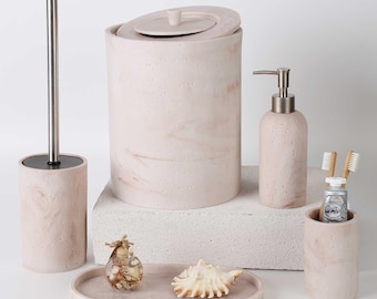 Op de afbeelding: Een badkameraccessoireset in een lichtbeige marmeren afwerking, inclusief een afvalemmer met deksel, een toiletborstel met metalen handvat, een zeepdispenser, een tandenborstelhouder en een ovaal dienblad met een glazen potje en een zeeschelp.