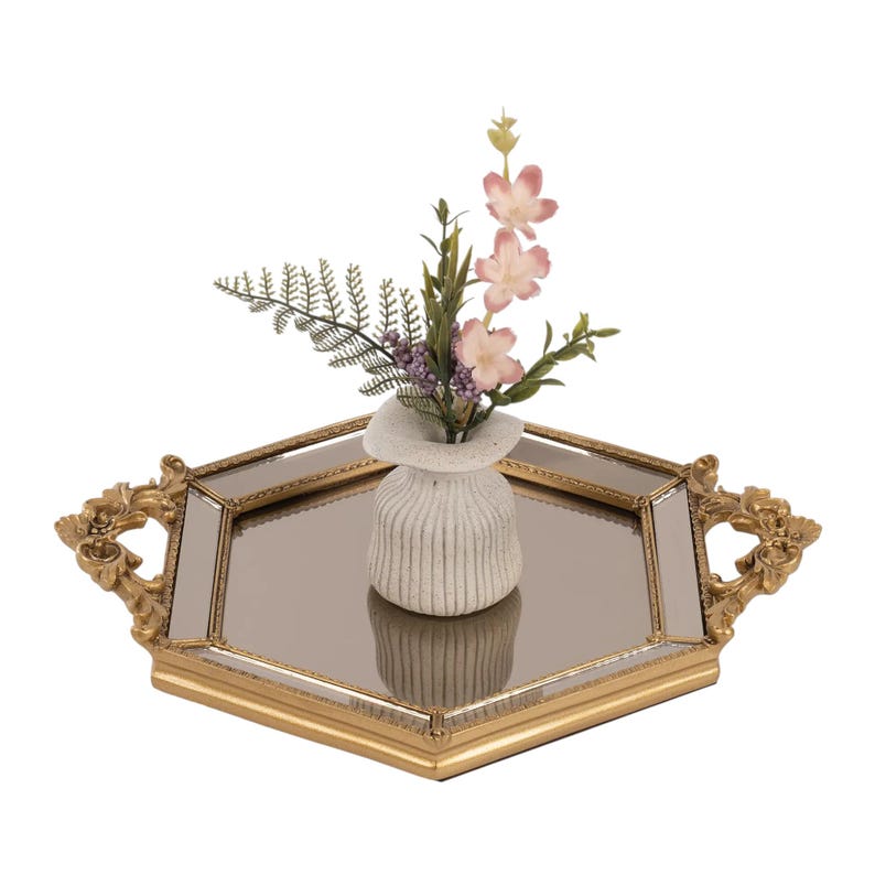 May include: A hexagonal mirrored tray with a gold-toned frame and ornate handles, featuring a small, ribbed, off-white ceramic vase placed in the centre. The vase contains a decorative arrangement of green fern leaves and light pink flowers.