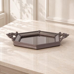 May include: A hexagonal mirrored tray with a dark metallic frame and ornate, decorative handles on two sides. The tray features a reflective glass base and a raised, textured border that surrounds the central mirror surface in a geometric shape.