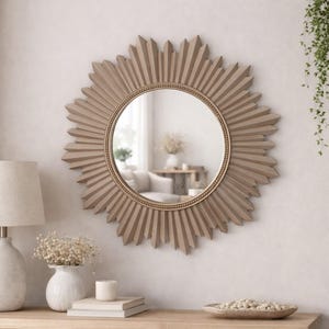Puede incluir: Un espejo de pared redondo con un marco en forma de sol que presenta listones de madera puntiagudos y radiantes en un acabado taupe mate. El centro de cristal circular está bordeado por un fino borde dorado con cuentas que crea contraste.
