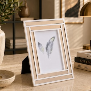 Puede incluir: Un marco de fotos rectangular con un diseño geométrico blanco y beige. El marco contiene una impresión de dos plumas grises y blancas. El marco está apoyado en un soporte negro. También se ven un jarrón con vegetación y un cuenco pequeño.
