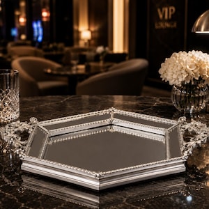 Può includere: Un vassoio a specchio argentato con manici decorati è appoggiato su una superficie scura e decorata. Un bicchiere di cristallo tagliato e un vaso di fiori bianchi sono nelle vicinanze. Lo sfondo presenta un bar scarsamente illuminato con un'insegna VIP lounge.