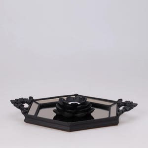 May include: A black decorative tray with a mirrored surface and a black floral candle holder. The tray has ornate handles on each side. The candle holder is in the shape of a flower.