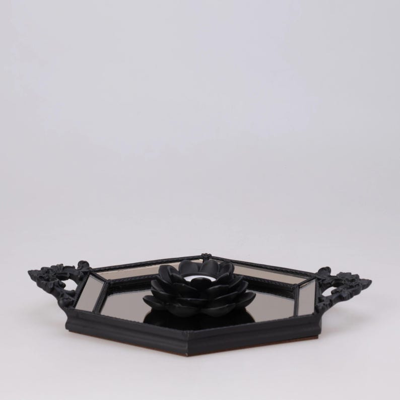 May include: A black decorative tray with a mirrored surface and a black floral candle holder. The tray has ornate handles on each side. The candle holder is in the shape of a flower.