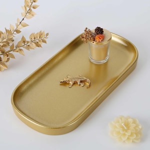 Puede incluir: Una bandeja ovalada de color dorado con una pequeña figura de caimán dorada en el centro. Un portavelas de cristal con una vela dorada y un pequeño arreglo floral seco se encuentra sobre la bandeja, junto a una vela de flor color crema.
