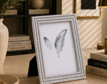 Puede incluir: Un marco de fotos rectangular con acabado plateado metálico y un borde texturizado que imita una cuerda. El marco contiene una lámina blanca con una ilustración impresa de dos plumas grises. Se mantiene en posición vertical con un soporte negro.