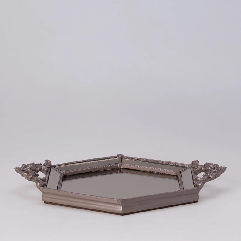 May include: A decorative, hexagonal tray with a mirrored surface and ornate handles. The tray is a muted silver color and has a raised edge. The handles feature a floral design.