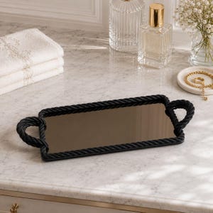 May include: A rectangular black tray with a mirrored surface and rope-style trim. The tray has two loop handles and is displayed on a marble countertop. The tray is surrounded by other bathroom accessories.