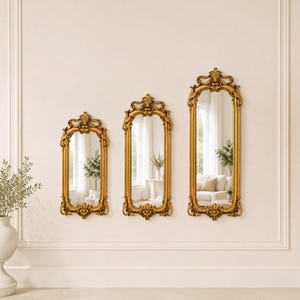 May include: Three vertical wall mirrors with ornate gold frames featuring intricate scrollwork and floral carvings at the top and bottom. The mirrors are arranged in a row, each with a different height, reflecting a bright room with furniture.