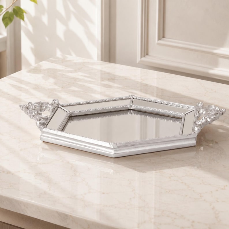 May include: A hexagonal mirrored tray with a silver-toned frame. The tray features a reflective glass base and decorative silver handles on two sides. The frame has a beaded edge detail, providing a structured border around the central mirror surface.