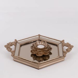 May include: A decorative gold-toned tray with a mirrored surface and ornate handles. A gold lotus-shaped candle holder with a lit white candle sits in the center. The tray has a hexagonal shape.