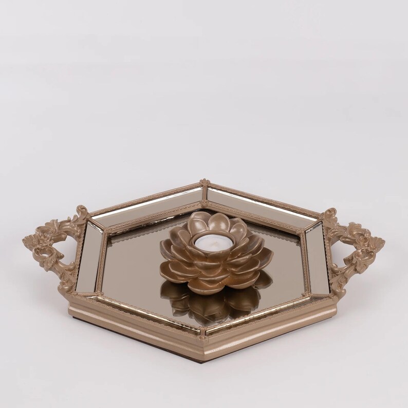 May include: A decorative gold-toned tray with a mirrored surface and ornate handles. A gold lotus-shaped candle holder with a lit white candle sits in the center. The tray has a hexagonal shape.