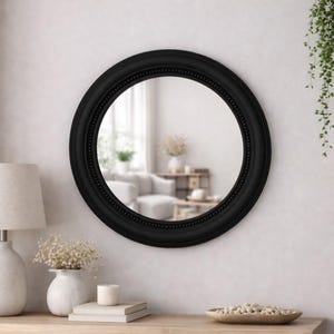 Pode incluir: Um espelho de parede redondo com uma moldura espessa em preto mate, apresentando uma textura subtil de contas ao longo da borda interna. A superf&iacute;cie circular de vidro reflete um espa&ccedil;o interior luminoso e desfocado.