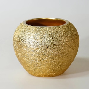 Puede incluir: Un jarrón dorado texturizado con forma redonda. El borde interior del jarrón es de un color dorado más oscuro. El exterior tiene una textura rugosa y granulada. El jarrón es probablemente de cerámica o un material similar.