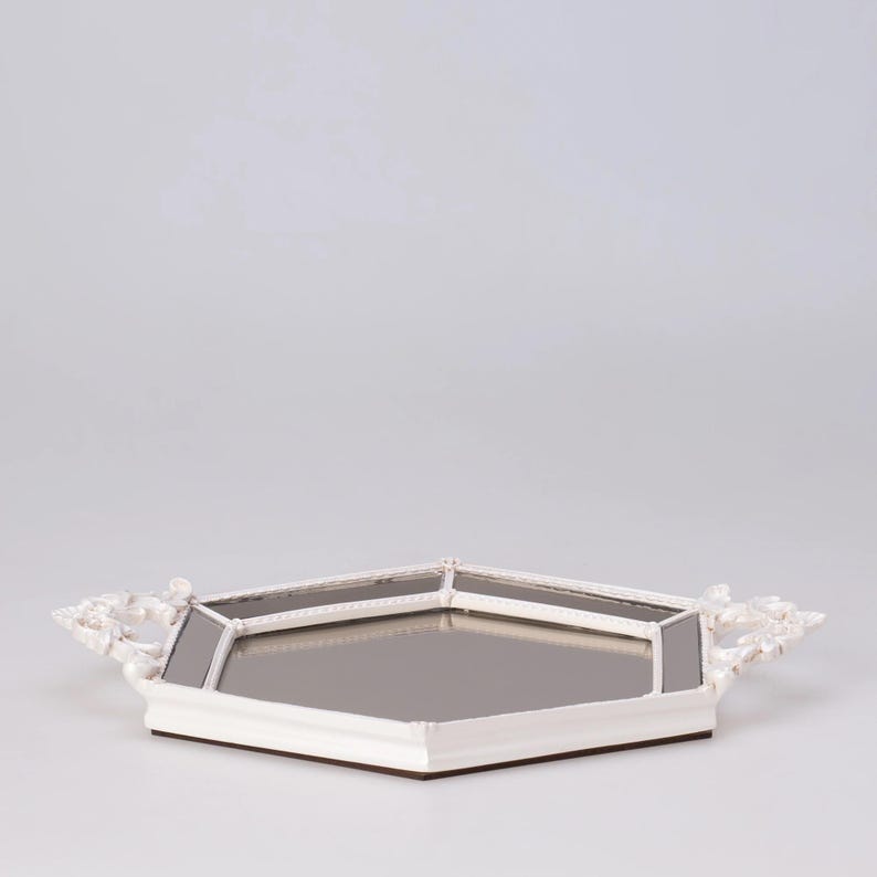 May include: A white, octagonal mirrored tray with ornate, floral-style handles. The tray has a mirrored surface and a white frame with a decorative trim. The base is a dark brown color.