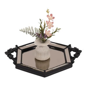 May include: A hexagonal mirrored tray with a black frame and ornate handles, featuring a small, off-white, ribbed ceramic vase in the centre. The vase contains a decorative arrangement of green fern leaves, small purple buds, and light pink flowers.