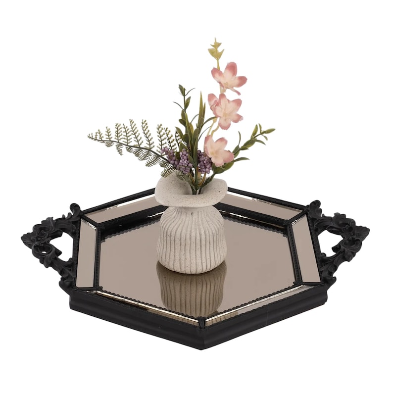 May include: A hexagonal mirrored tray with a black frame and ornate handles, featuring a small, off-white, ribbed ceramic vase in the centre. The vase contains a decorative arrangement of green fern leaves, small purple buds, and light pink flowers.