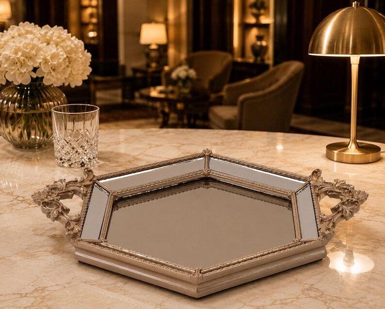 May include: A decorative, mirrored tray with a light beige frame and ornate handles. The tray has a geometric shape and is placed on a marble surface. A vase of white flowers, a glass, and a gold lamp are also visible.
