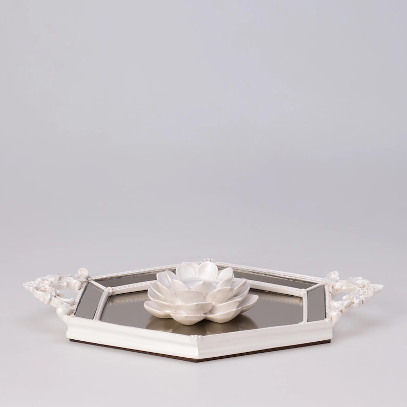 May include: A white, octagonal decorative tray with a mirrored surface and a white ceramic lotus flower in the center. The tray has ornate, floral-patterned handles on each side. The tray is a decorative home accent.