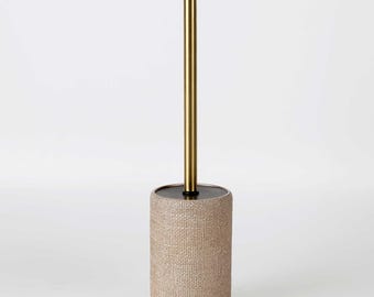 May include: A toilet brush set featuring a cylindrical holder with a textured beige woven exterior and a smooth black top lid. The long, straight handle is made of polished gold-toned metal, extending vertically from the centre of the holder.