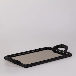 Op de afbeelding: Een rechthoekig zwart dienblad met een touwachtige rand en een spiegeloppervlak. De lade heeft een handvat van hetzelfde touwmateriaal. Het ontwerp is modern en minimalistisch, geschikt voor serveren of presenteren.