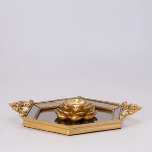 May include: A gold-colored decorative tray with a mirrored surface and a gold lotus flower candle holder. The tray has ornate handles and a geometric shape, perfect for home decor.