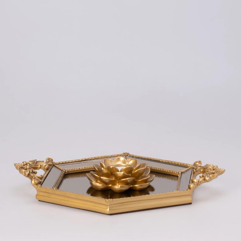 May include: A gold-colored decorative tray with a mirrored surface and a gold lotus flower candle holder. The tray has ornate handles and a geometric shape, perfect for home decor.