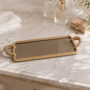 May include: A rectangular gold-toned tray with a mirrored surface. The tray is framed with a rope-like design and features looped handles at each end. The tray is likely used for decorative purposes.