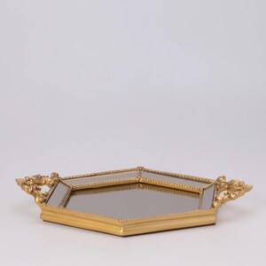 May include: A gold-colored, geometric tray with a mirrored surface. The tray has ornate handles on each end. The tray is set against a plain white background. The tray is likely used for decorative purposes.