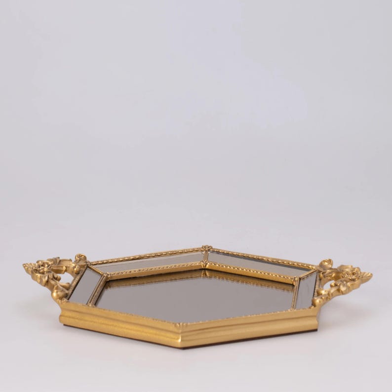 May include: A gold-colored, geometric tray with a mirrored surface. The tray has ornate handles on each end. The tray is set against a plain white background. The tray is likely used for decorative purposes.