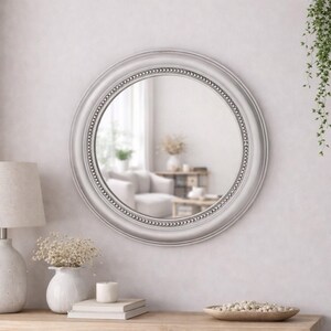 Pode incluir: Um espelho de parede redondo com uma moldura lisa cinza-clara, apresentando uma borda interna decorativa com contas. A superf&iacute;cie circular de vidro reflete um espa&ccedil;o interior luminoso e desfocado, capturando a luz e a profundidade do ambiente.